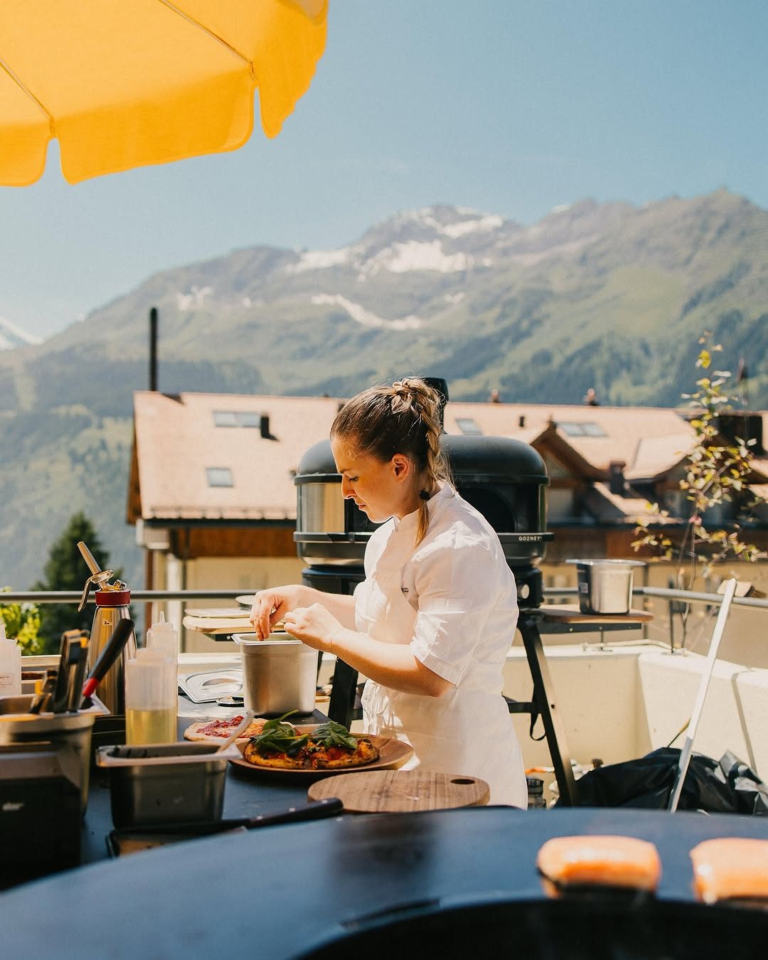 A cook at the wood-fired grill on the terrace, mountains behind