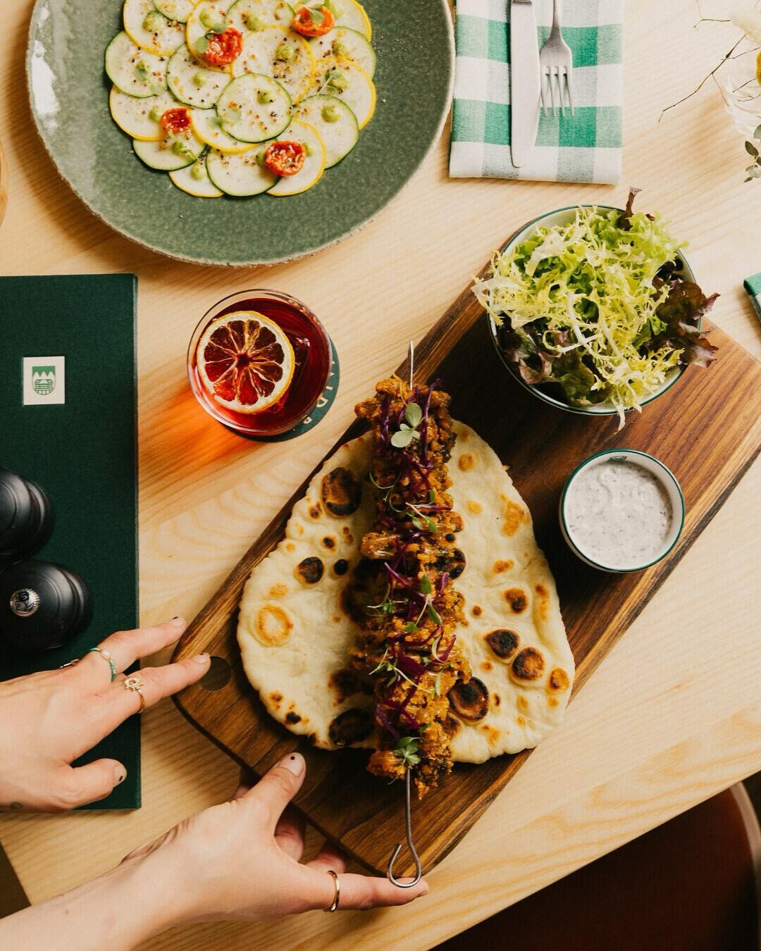 Shared dishes on a wooden table seen from above: a courgette carpaccio, a skewer on flatbread, a salad and a negroni